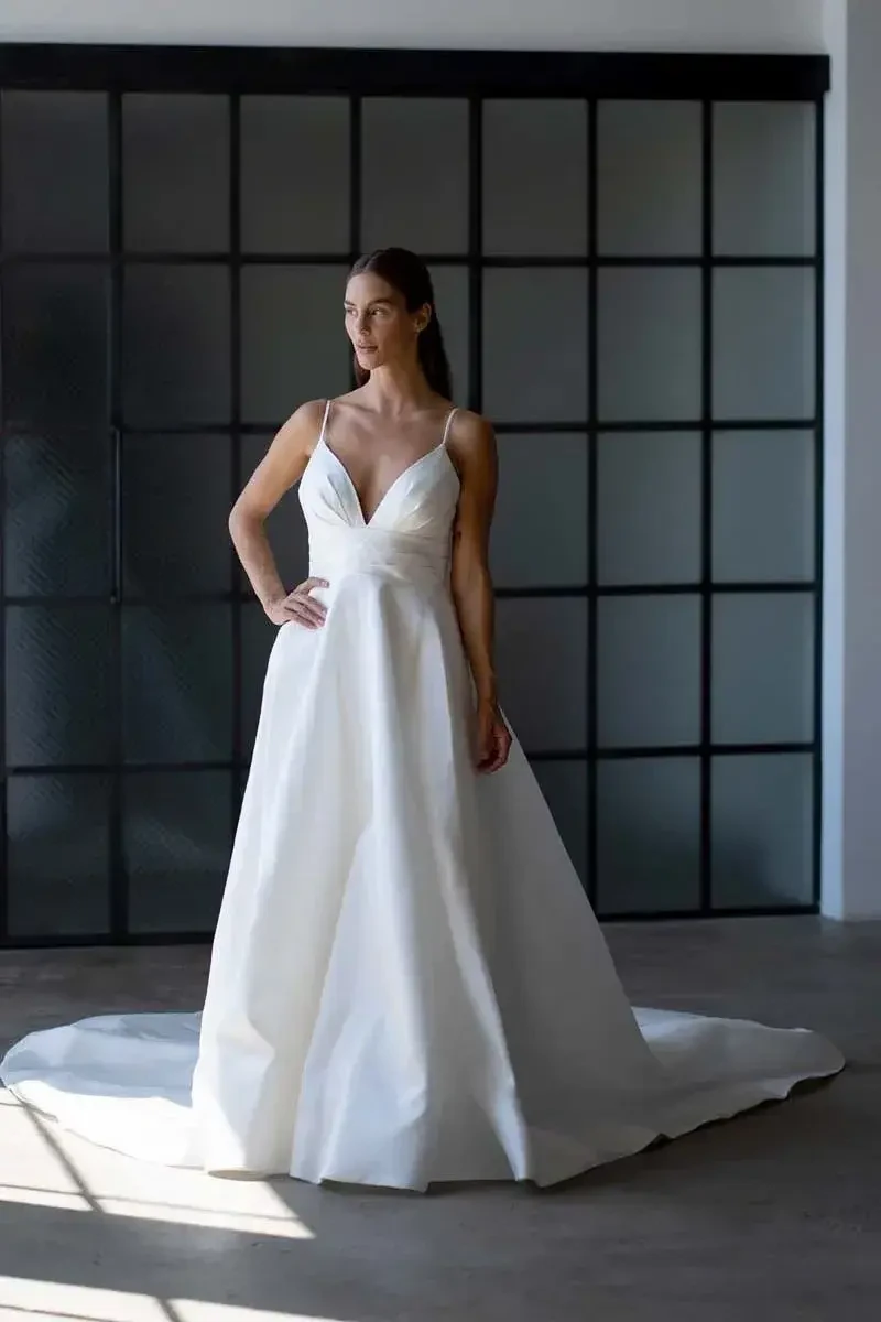 A woman wearing a white bridal gown with a fitted bodice and flowing skirt, standing in a spacious room with large windows.