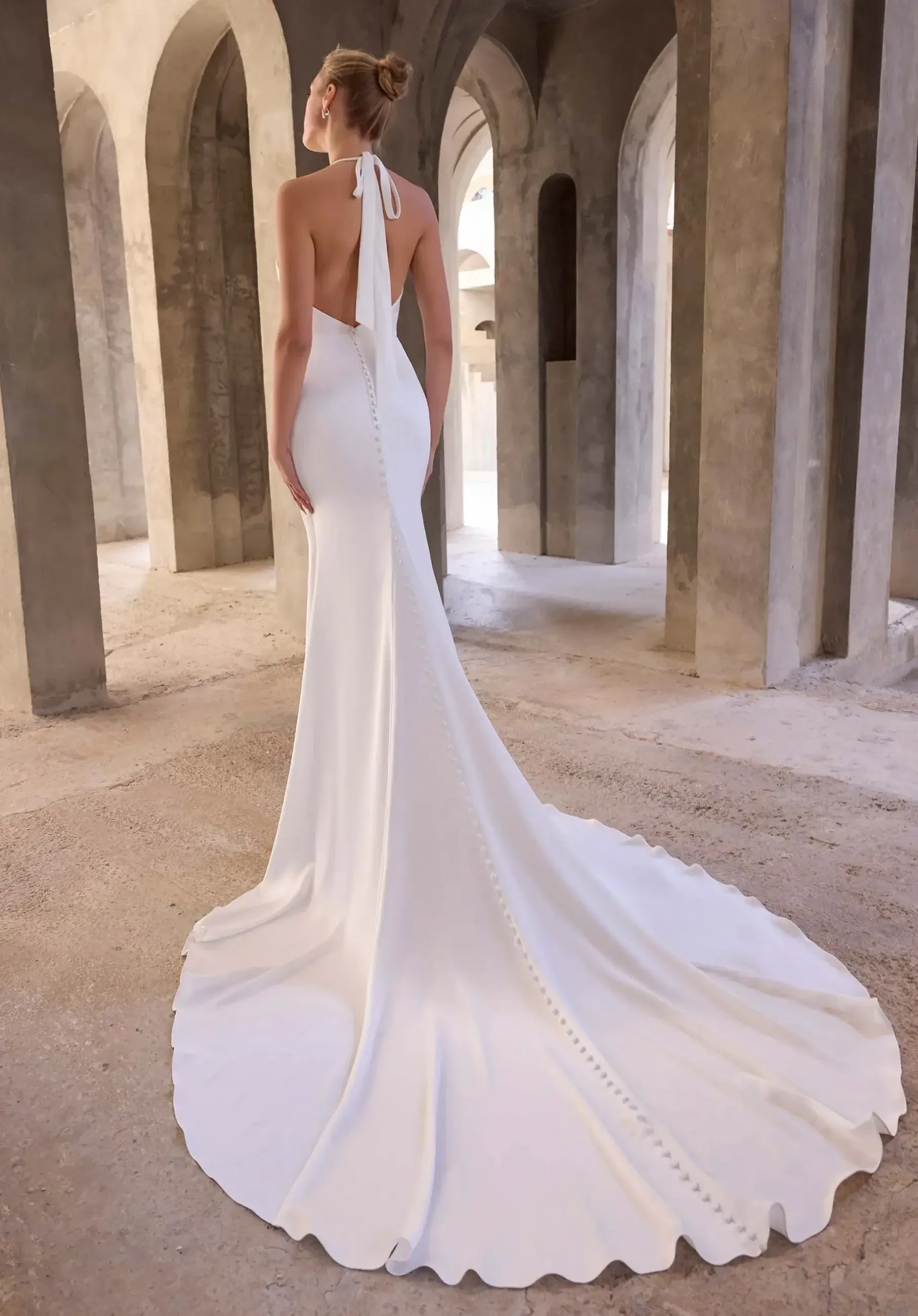 A woman in a sleek, backless white gown with a long train stands in an arched stone hallway, evoking elegance and a sense of timelessness.