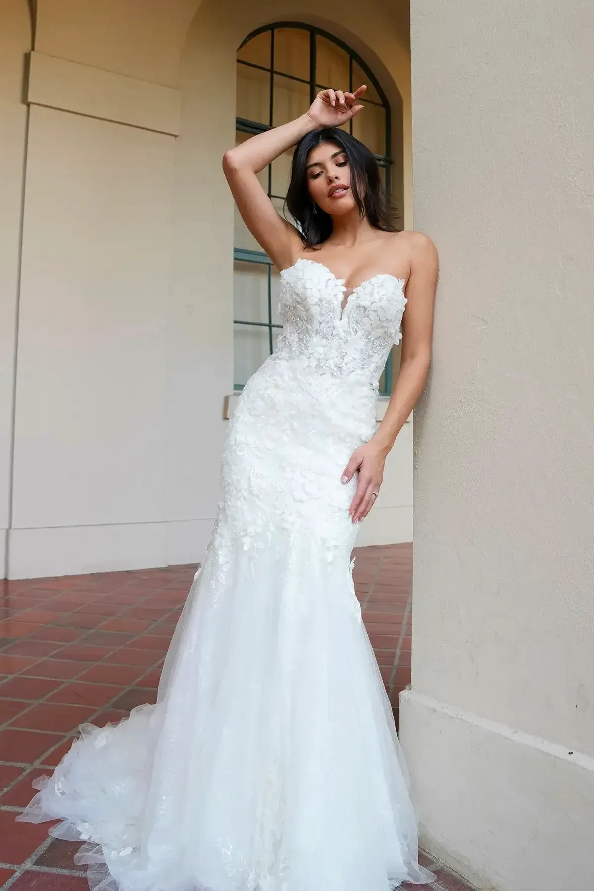 A model poses in a fitted white wedding dress with lace details, leaning against a wall in a well-lit indoor setting.