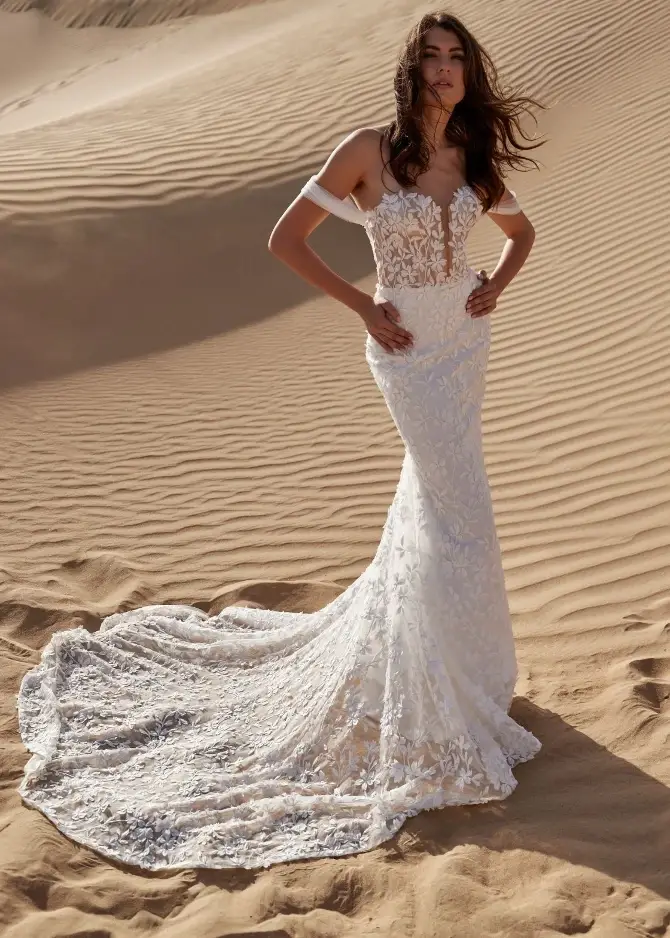A model wearing a fitted wedding gown with lace details, posing on sand dunes.