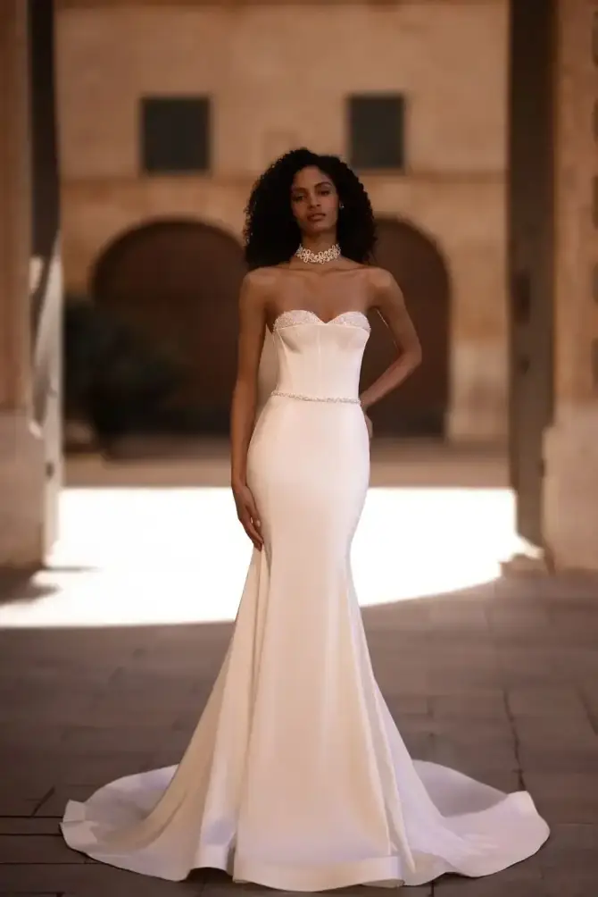 Elegant woman in a fitted, strapless white bridal gown stands confidently in a sunlit archway. The setting is warm and inviting, conveying grace.