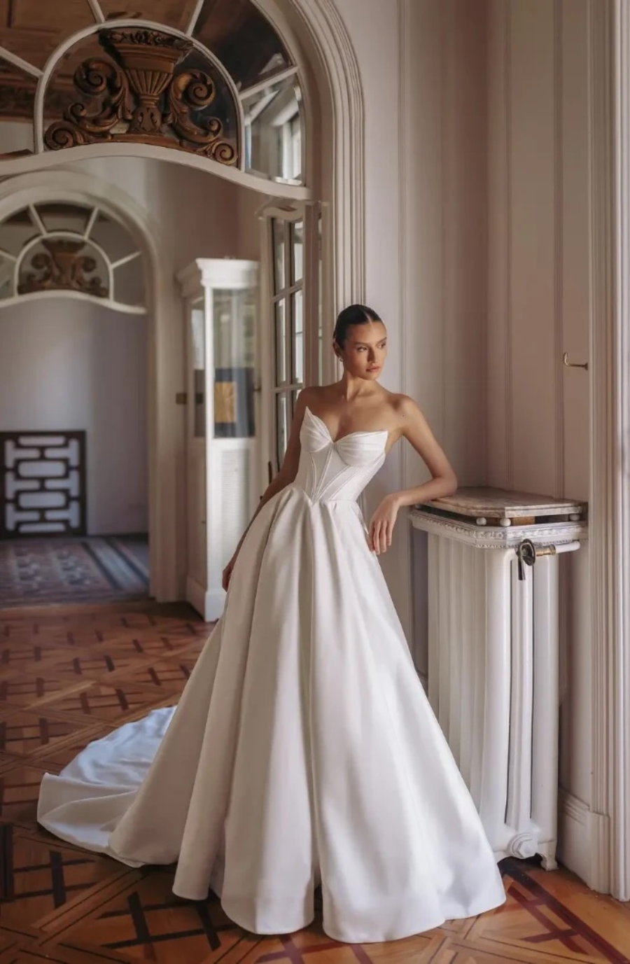 A woman in an elegant white wedding dress with a sweetheart neckline leans against a radiator in a grand hallway. The space has arched doorways and a warm, refined ambiance.
